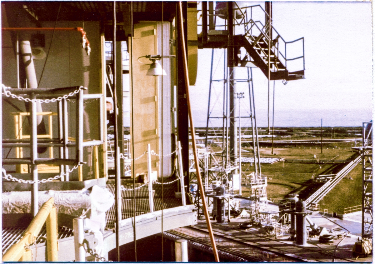 Image 025. From the lowest main floor level of the Rotating Service Structure at Launch Complex 39-B, Kennedy Space Center, Florida, we find ourselves looking across toward the northeast, at the fuel side of the elevation 112’-0” floor grating, APU/APS access platforms, Orbiter Mold Line framing cutout with removable handrail posts and safety chains, and the APU Valve Complex Access Doors, with the Hinged Tail Service Mast Access Stair hanging out over empty space above and to the right, with a slice of the Flame Trench and Pad Deck in view, and beyond that the Sound Suppression System Water Tower, with the LH2 Burn Pond as a lighter-shaded rectangular area in the grass beyond it, and to the right of that, the cross-country cryo line which facilitated Liquid Hydrogen fill and drain operations from the very large Dewar it was stored in, a tiny bit of which can be seen in the distance on the far right margin of the photograph, behind the framing steel which makes up the top of the MLP East Stair Tower, also only partly visible on the far right margin. Photo by James MacLaren.